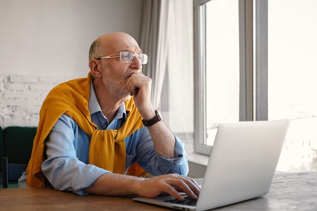 Foto van aantrekkelijke stijlvolle zeventig-jarige senior zakenman dragen bril en formele kleding met doordachte peinzende blik tijdens het werken op laptop pc, zittend aan een bureau bij raam