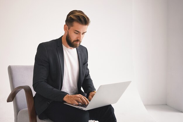 Foto van aantrekkelijke succesvolle jonge Europese bebaarde mannelijke ondernemer die op afstand werkt, e-mail controleert op draagbare computer, ernstige gezichtsuitdrukking heeft, gericht op zakelijke kwesties