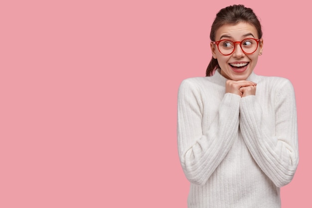 Foto van aantrekkelijke vrouw glimlacht breed, houdt de handen onder de kin, tevreden met een goed moment in het leven, draagt een casual witte trui