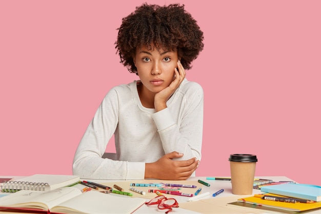 Foto van aantrekkelijke zwarte ontwerper met Afro kapsel, zit op de werkplek, draagt een witte trui, maakt tekeningen met kleurpotloden, geïsoleerd over roze muur, geniet van koffie