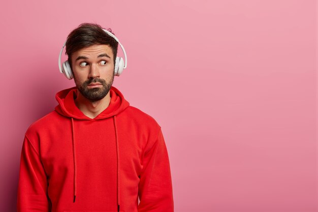 Foto van bebaarde hipster-man kijkt weg met verbaasde verwonderde blik, gekleed in rood sweatshirt, ziet iets ongelooflijks, gebruikt koptelefoon, geïsoleerd over roze pastel muur, kopieer ruimte opzij