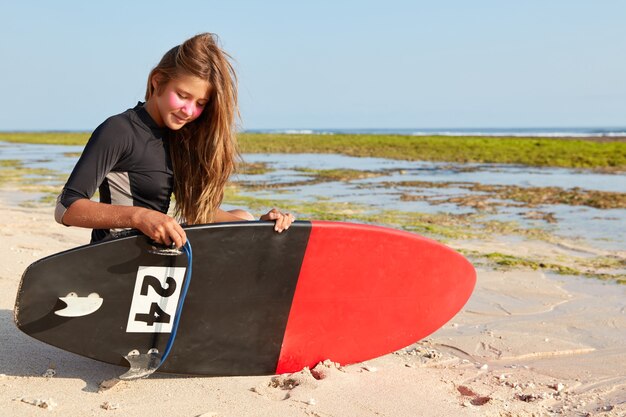 Foto van beginnende vrouwelijke surfer probeert board te repareren, gekleed in een zwart wetsuit, heeft een zink surfmasker op het gezicht