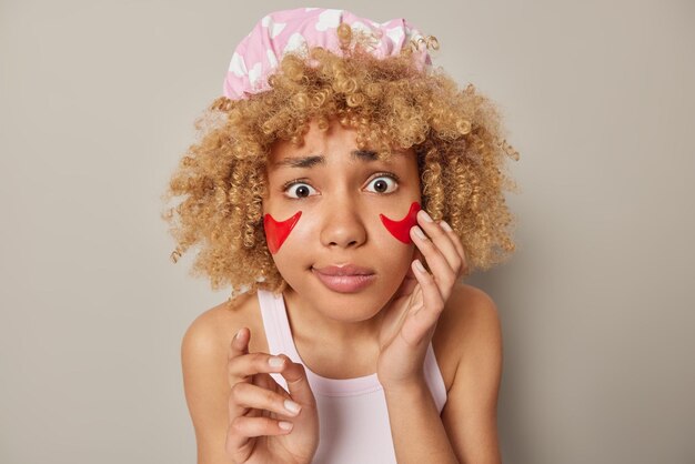 Foto van bezorgde vrouw met krullend haar draagt een badmuts en een casual t-shirt brengt rode schoonheidspleisters onder de ogen aan en zorgt voor de gevoelige huid geïsoleerd over grijze achtergrond Cosmetologiebehandelingen