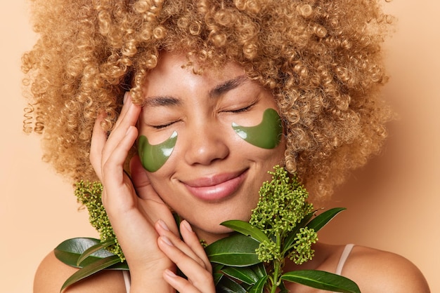 Gratis foto foto van blije jonge vrouw met krullend borstelig haar houdt hand op gezicht glimlacht gelukkig heeft ogen gesloten houdt groene plant past schoonheidspleisters onder ogen gebruikt natuurlijke cosmetica huidverzorgingsconcept