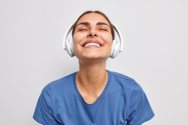 Foto van blije tevreden vrouw houdt ogen gesloten van plezier vangt elk stukje nummer luistert muziek via draadloze koptelefoon lacht tandjes draagt casual blauw t-shirt poseert tegen witte muur
