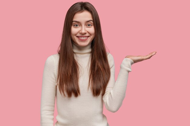 Foto van blije vrouwelijke verkoper werpt palm boven roze lege ruimte, gekleed in vrijetijdskleding, glimlacht zachtjes, adverteert nieuw item
