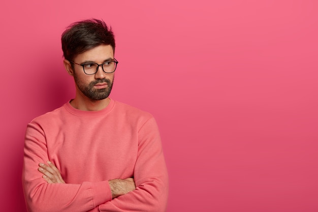 Foto van contemplatieve ongeschoren man met bril houdt de handen gekruist over de borst, denkt na over het voorbereiden van iets interessants voor een project, overdenkt hoe de situatie kan worden opgelost, gekleed in een roze trui