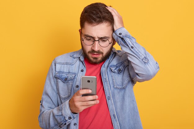 Foto van de charismatische geschokte jonge gebaarde mens die zijn smartphone houdt, aandachtig bekijkend het scherm van het apparaat, hand op het hoofd zet