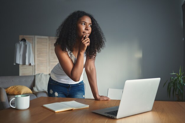 Foto van doordachte donkere student meisje in tanktop en spijkerbroek kin aanraken met peinzende gezichtsuitdrukking, pen vasthouden tijdens het schrijven van essay, huiswerk doen, staande voor open laptop