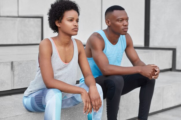 Foto van doordachte sportieve donkere vrouw en man rust na oefeningen op trappen, motivatie om gezond en fit te blijven, gekleed in actieve kleding, samen trainen. Atletische trainingen