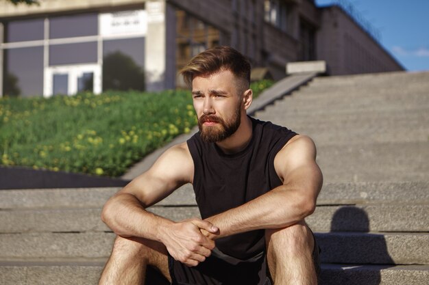 Foto van doordachte uitgeputte jonge mannelijke atleet met pluizige baard en gebruinde huid zittend op betonnen treden, ontspannen na intensieve cardiotraining sport, fitness, stijl en mode-concept