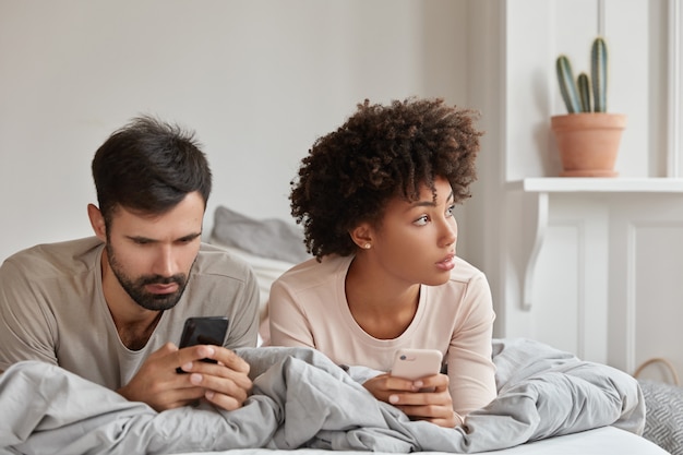 Foto van doordachte verslaafde vrouw en man van gemengd ras lezen website via mobiele telefoontoepassing, tijd doorbrengen in bed op zaterdagochtend, hebben technologieverslaving. Paar surfen op internet