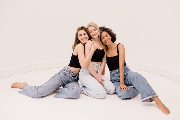 Foto van drie mooie multi-etnische vrouwen in vrijetijdskleding die bij elkaar zitten en glimlachen naar de camera op witte achtergrond