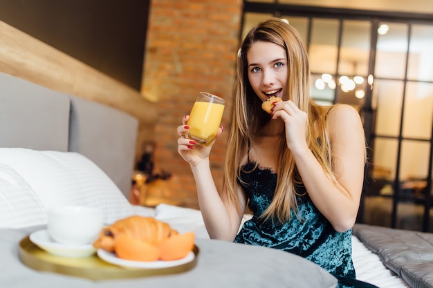 Foto van een gelukkige blonde vrouw ontbijten op slaapkamer met verse jus d'orange en croissant