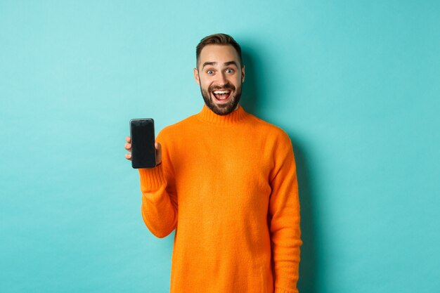 Foto van een gelukkige man die mobiel scherm toont, online winkel, applicatie introduceert, staande over turkooizen muur.