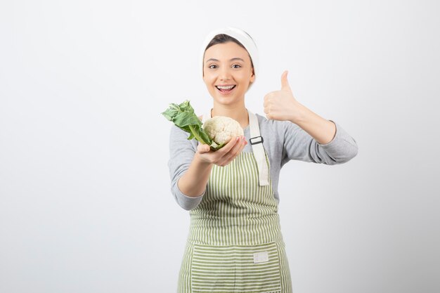 Foto van een jong aardig vrouwenmodel in schort met een bloemkool die een duim omhoog toont