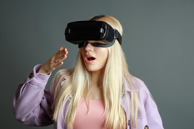 Foto van een jonge geschokte blonde met een VR-bril op een grijze achtergrond