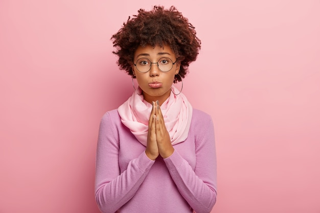 Gratis foto foto van een jonge vrouw met een donkere huid en afro-kapsel, heeft een zielige uitdrukking, houdt de handpalmen tegen elkaar gedrukt, vraagt om nog een kans, gelooft in beter, draagt een ronde bril, paarse trui