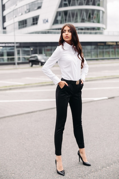Foto van een knappe blanke vrouw met lang donker golvend haar in een wit overhemd, zwarte broek en hakken poseren voor de camera