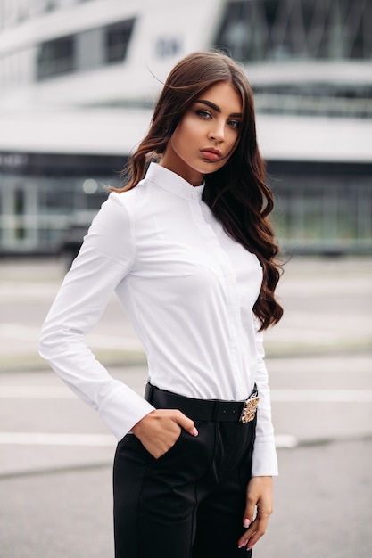 Foto van een knappe blanke vrouw met lang donker golvend haar in een wit overhemd, zwarte broek en hakken poseren voor de camera