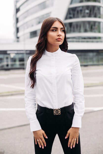 Foto van een knappe blanke vrouw met lang donker golvend haar in een wit overhemd, zwarte broek en hakken poseren voor de camera