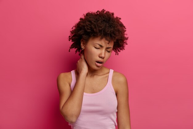Foto van een noodlijdende vrouw raakt nek, lijdt aan rug- of wervelkolomziekte, gaapt en sluit de ogen, heeft een vermoeide uitdrukking, nonchalant gekleed, heeft pijnlijke gevoelens, geïsoleerd over roze muur