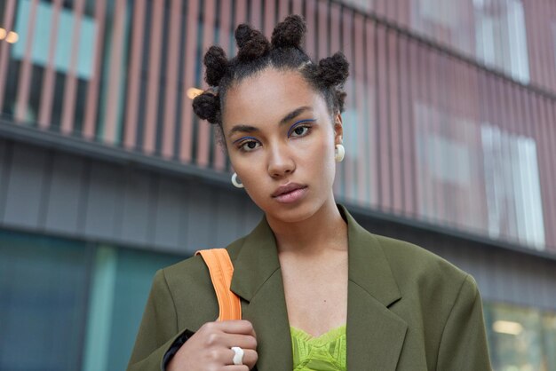 Foto van een serieus hipstermeisje met haarknotjes blauwe eyeliner gekleed in een formeel donkergroen jasje kijkt direct naar de camera loopt naar buiten poses luistert aandachtig naar iets