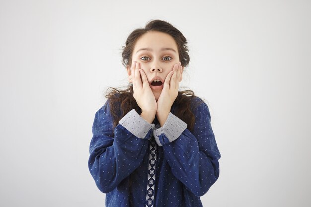 Foto van een verbaasd gefascineerd meisje met donker haar dat de mond wijd openzet, de handen op haar wangen vasthoudt, volledig ongeloof en geschokt uitdrukt en onverwacht verbazingwekkend nieuws ontvangt. Echte emoties