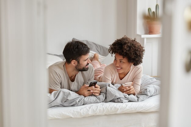 Foto van een vrolijke vriendin en vriend zoeken een hotel om te verblijven tijdens de vakantie, blader door de applicatie op de smartphone, chat met vrienden, geniet van comfort in de slaapkamer. Moderne technologieën concept