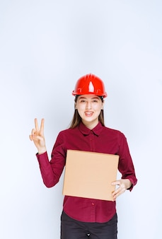 Foto van een vrouw in een rode helm die een kartonnen doos vasthoudt en een overwinningsteken geeft.