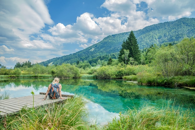 Foto van een vrouw zittend op een houten brug tegen een smaragdgroen meer met een adembenemende natuur