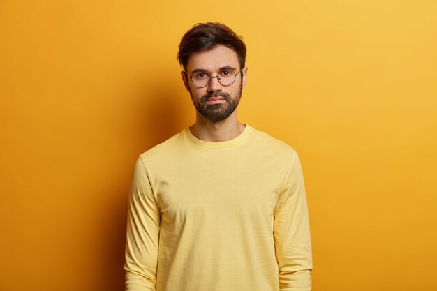 Foto van ernstig uitziende man heeft donkere haren, draagt een ronde bril en gele trui, directe blik, poseert binnen, heeft een informeel gesprek met iemand. Monochroom. Gezichtsuitdrukkingen concept