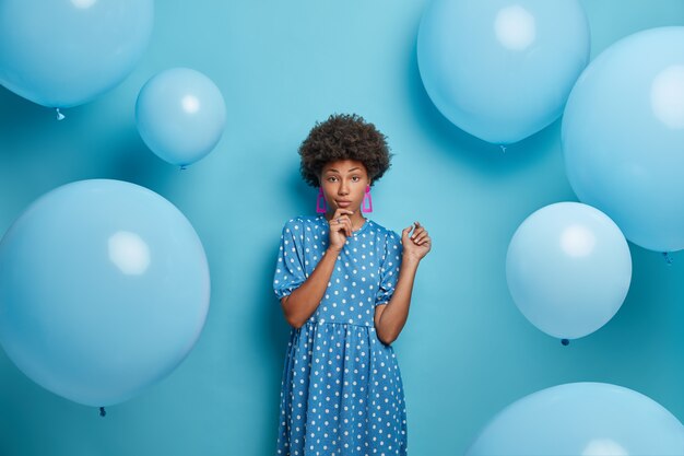 Foto van ernstige vrouw met krullend haar, gekleed in modieuze kleding, geniet van feest, vormt tegen blauwe muur, heeft een aangenaam gesprek. Mooie dame viert verjaardag, heeft een geweldige dag