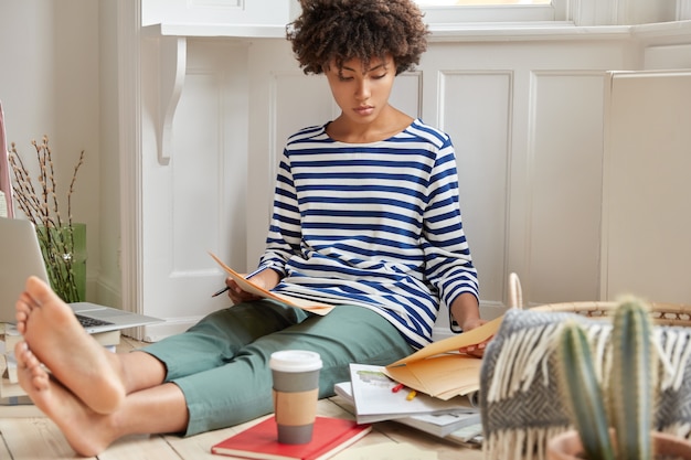 Foto van ernstige zwarte jonge vrouw analyseert contract, aromatische koffie drinkt, heeft een attente blik