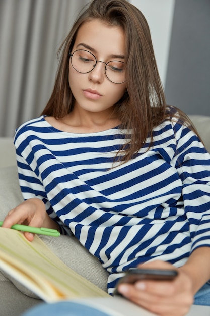 Foto van geconcentreerde vrouwelijke lezer leest boek, onderstreept informatie met pen, probeert haar vocabulaire te verrijken, houdt moderne mobiele telefoon vast, draagt optische bril voor goed zicht, heeft een serieuze blik