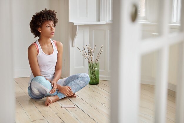 Foto van geconcentreerde zwarte vrouw zit gekruiste benen op houten vloer