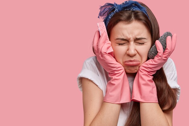 Foto van gefrustreerde drukke jonge vrouw fronst gezicht in ontevredenheid, houdt handen in de buurt van gezicht, reinigt appartement