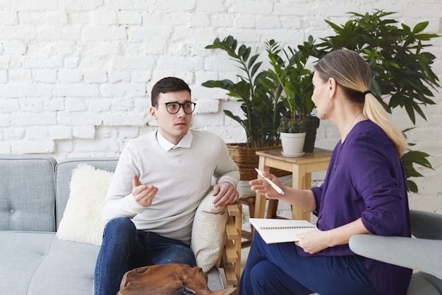 Foto van gefrustreerde jonge blanke man met trui en bril zittend op een comfortabele bank, zijn persoonlijke problemen delen met vrouwelijke counselor van middelbare leeftijd tijdens therapiesessie
