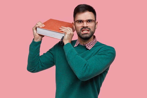 Foto van geïrriteerde Europese ongeschoren man houdt dikke rode oude encyclopedie vast, voelt zich geïrriteerd door het leren van nieuw materiaal