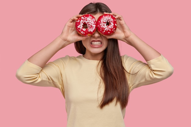 Foto van geïrriteerde jonge vrouw klemt tanden met woede, houdt smakelijke donuts op de ogen, heeft een aangename uitstraling, gekleed in vrijetijdskleding