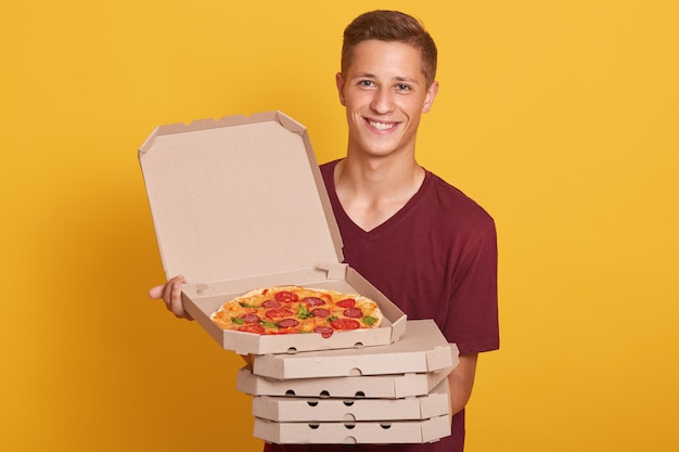Gratis foto foto van gelukkige blije mens die de toevallige t-shirt van bourgondië draagt, glimlachend camera bekijkt en pizzadozen houdt