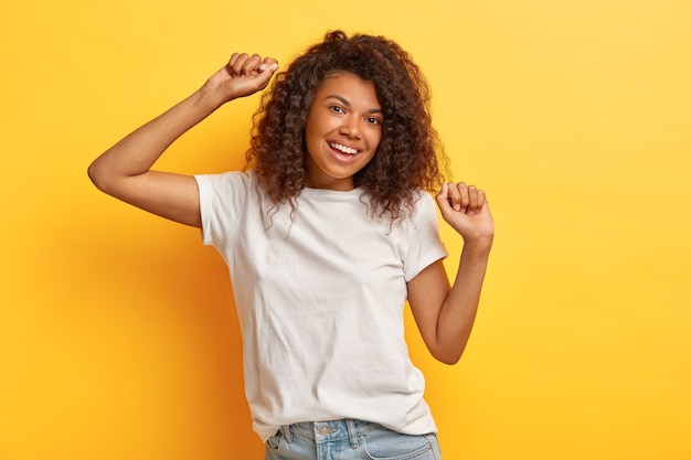 Foto van gelukkige donkerharige vrouw met positieve uitdrukking, werpt armen en beweegt tijdens het dansen, gekleed in wit casual t-shirt en spijkerbroek