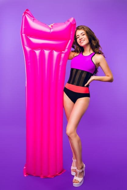 Foto van gemiddelde lengte van aantrekkelijk meisje met lang krullend haar poseren voor camera op paarse achtergrond in de studio. Ze draagt een badpak en houdt een roze luchtbed in de buurt.