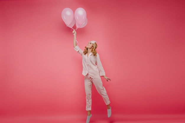 Foto van gemiddelde lengte van het enthousiaste blondemeisje poseren met ballonnen. Indoor portret van vrolijke dame in slaapmasker staande op haar tenen in haar verjaardag.