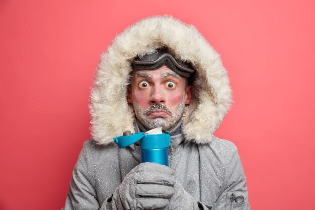 Gratis foto foto van geschokt beschaamd man beeft van kou besteedt veel tijd buiten tijdens ijzig weer, dranken hete thee houdt thermosfles draagt outfit voor wintersport.