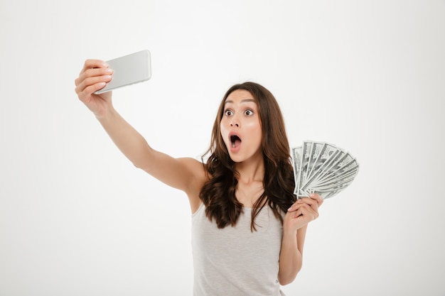 Foto van geschokte grappige vrouw die selfie fotograferen op zilveren mobiel, telefoon maken terwijl het houden van ventilator van dollarrekeningen over witte muur wordt geïsoleerd