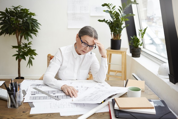 Foto van gestrest boos middelbare leeftijd vrouwelijke ingenieur dragen wit overhemd en bril kijken naar blauwdrukken of projectdocumentatie voor haar op bureau, gefrustreerd om zoveel fouten te zien