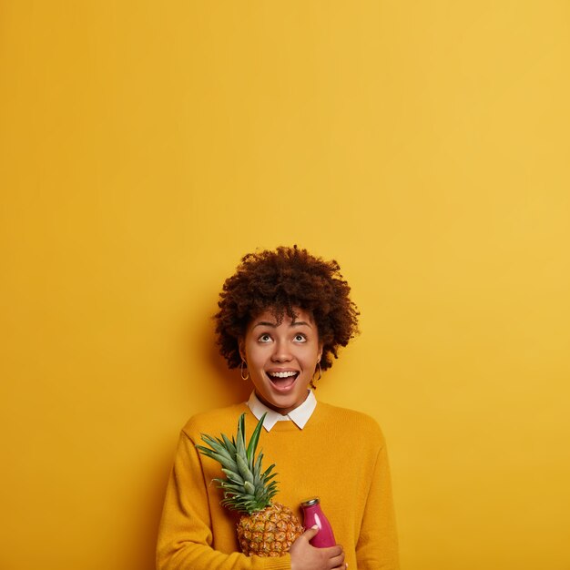 Foto van grappige etnische vrouw naar boven geconcentreerd, houdt verse ananas en smoothie, heeft gezonde voeding, gekleed in vrijetijdskleding, geïsoleerd over felgele muur, kopieer ruimte voor uw tekst