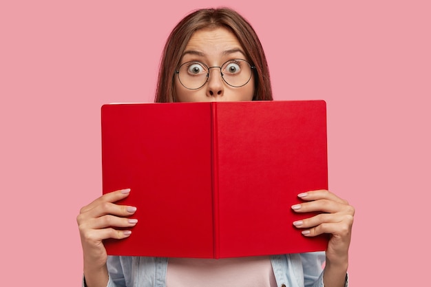 Foto van intelligente vrouwelijke student bedekt gezicht met rood boek, staart met afgeluisterde ogen, voelt geschokt om plotseling nieuws te horen, propt voor examen op de universiteit, geïsoleerd over roze muur