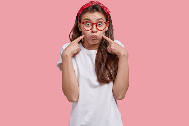 Foto van jonge blanke vrouw blaast wangen, maakt grimas, wijst naar het gezicht, draagt hoofdband, casual wit t-shirt